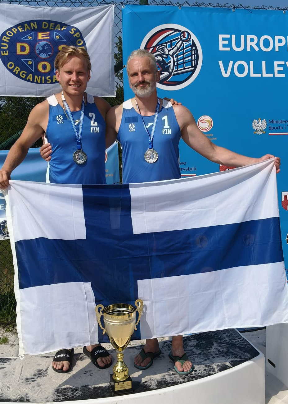 Ropponen ja Ahola levittävät Suomen lippua mitalit kaulassaan kameralle hymyillen. Lipun edessä maassa iso kultainen pokaali.